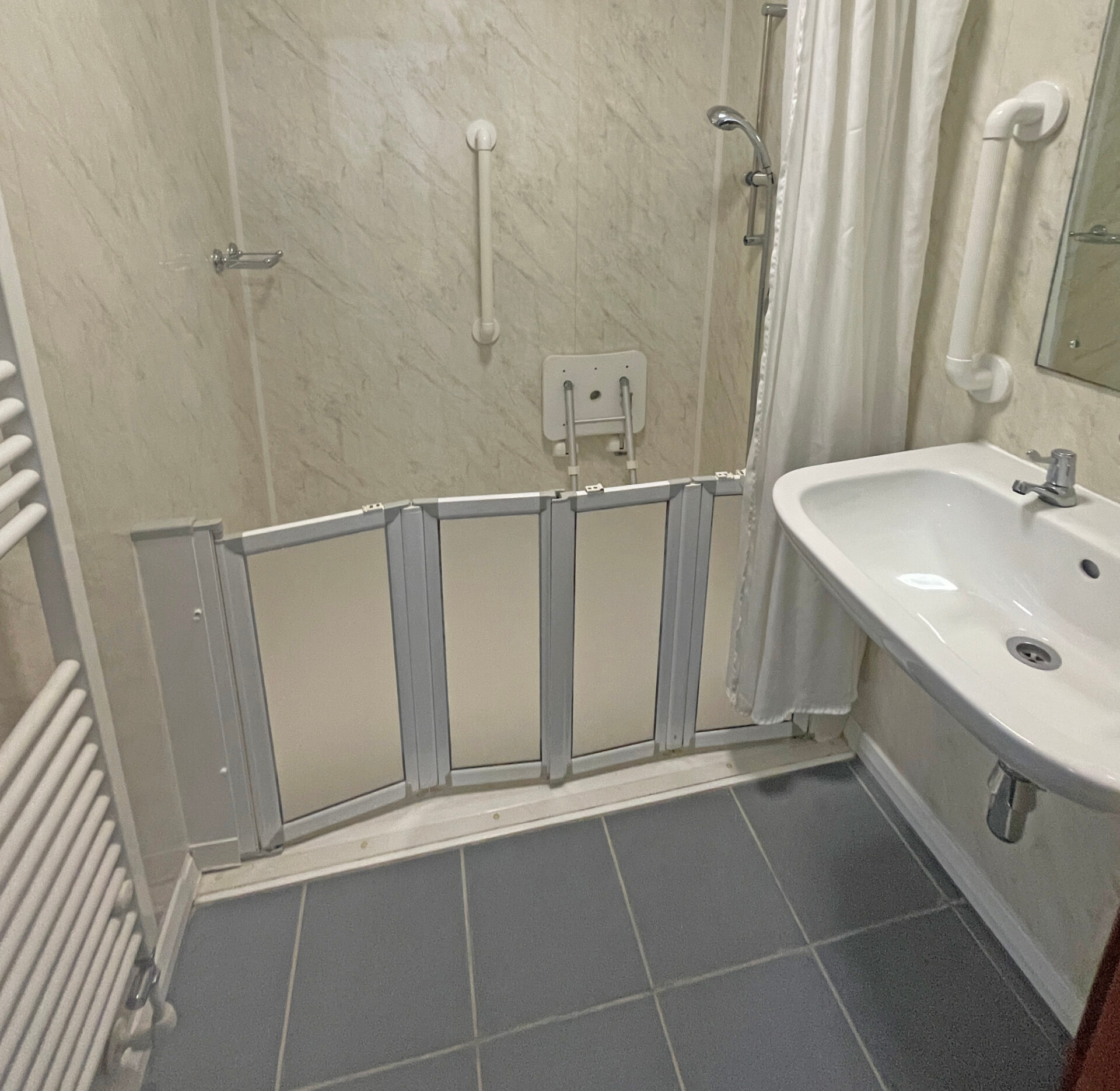 Wet Room3 A spacious wet room with white and grey tiled walls, a non-slip floor, and a walk-in shower area. The shower has a handheld and overhead fixture, with a grab rail mounted on the wall. A white fold-down shower seat is attached to the wall for accessibility. To the right, there is a white sink with a mirror above it, and a towel rail mounted nearby. The room is well-lit and designed for accessibility and ease of use.